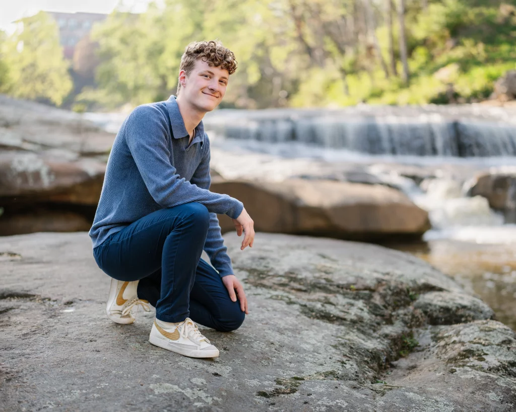 Jacobi's senior photos taken at Falls Park, Greenville SC: Young person with curly hair wearing a blue sweater, blue pants, and white sneakers, kneeling on a large rock near a flowing waterfall in a natural outdoor setting.