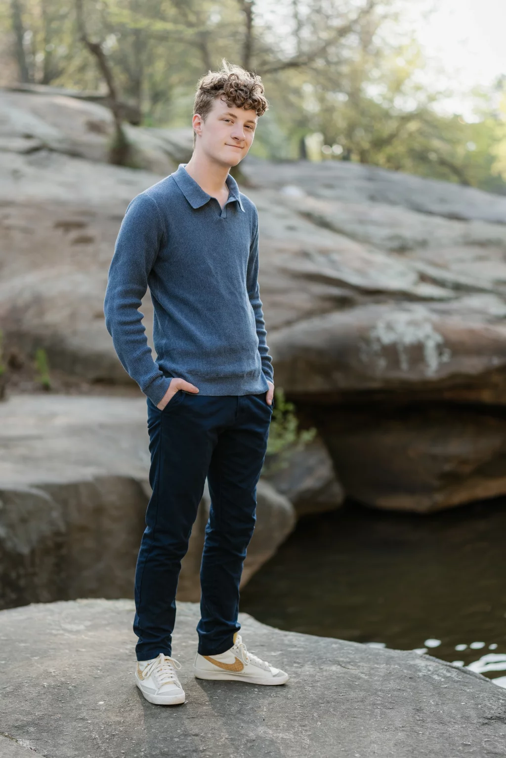 Jacobi's senior photos taken at Falls Park, Greenville SC: A young man with curly hair stands on a large rock near water, wearing a blue sweater, dark pants, and white sneakers, with a background of rocks and trees.