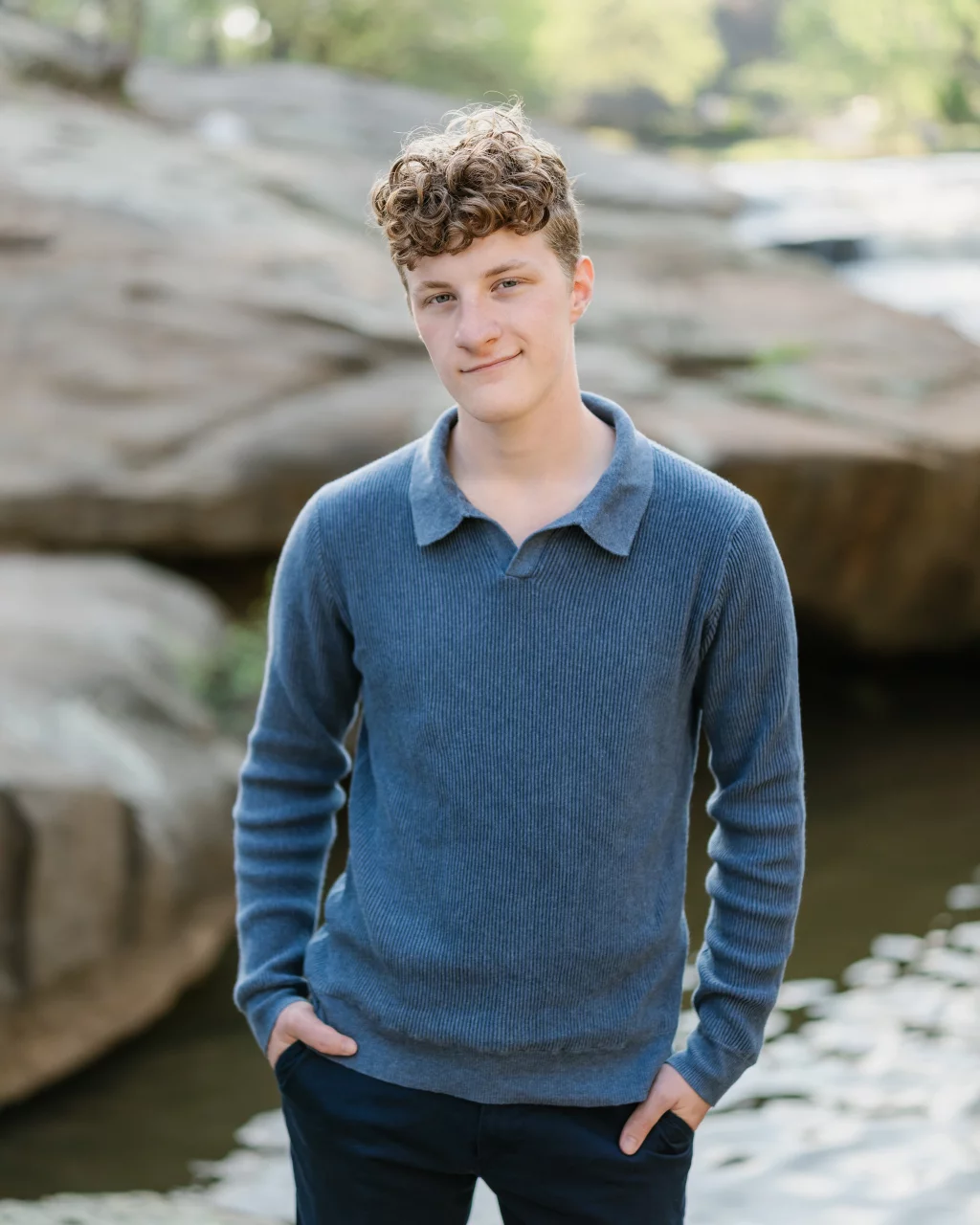Jacobi's senior photos taken at Falls Park, Greenville SC: Teenage boy with curly hair wearing a blue sweater, standing outdoors with hands in pockets in front of large rocks and water.