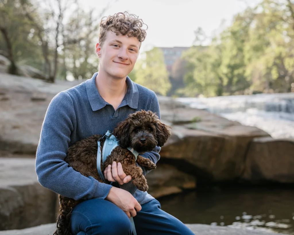 Jacobi's senior photos taken at Falls Park, Greenville SC: A young man with curly hair wearing a blue sweater sits outdoors by a rocky stream, smiling and holding a small brown dog in a harness.