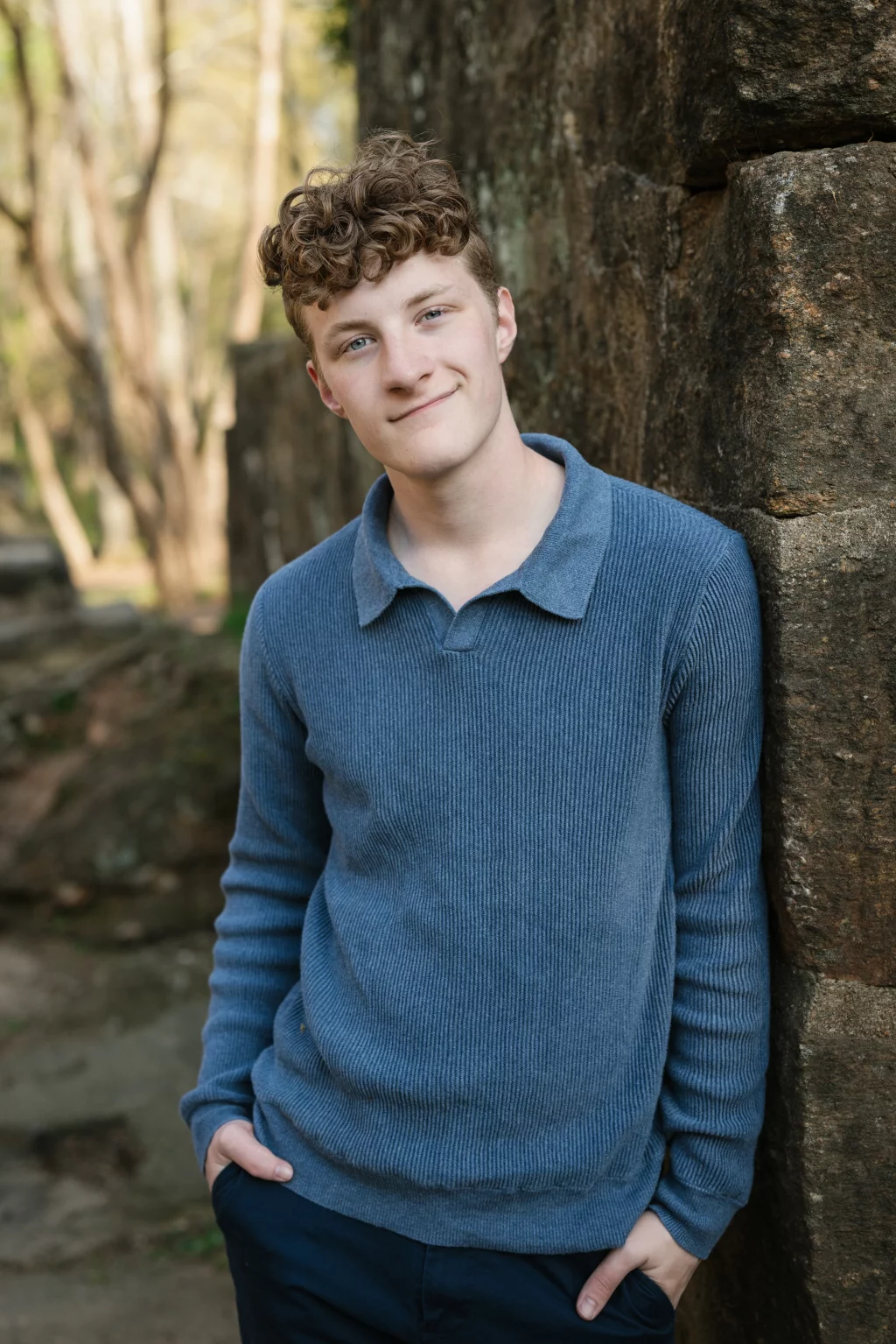 Jacobi's senior photos taken at Falls Park, Greenville SC: Young person with short curly hair wearing a blue sweater, standing outdoors and leaning against a stone wall, with hands in pockets and trees in the background.
