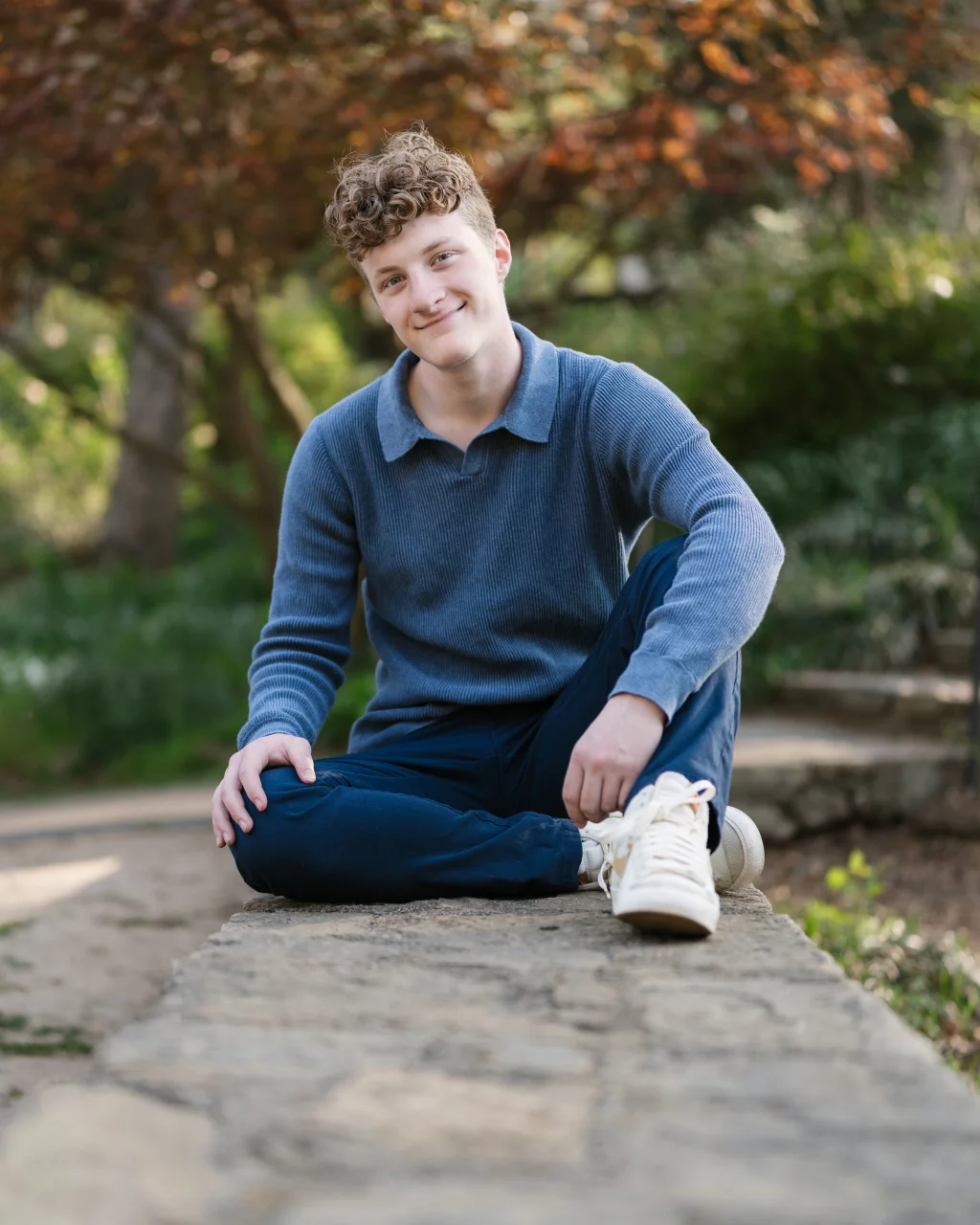 Jacobi's senior photos taken at Falls Park, Greenville SC: Young person with curly hair wearing a blue sweater and white sneakers, sitting cross-legged on a stone ledge in a park with green foliage and trees in the background.