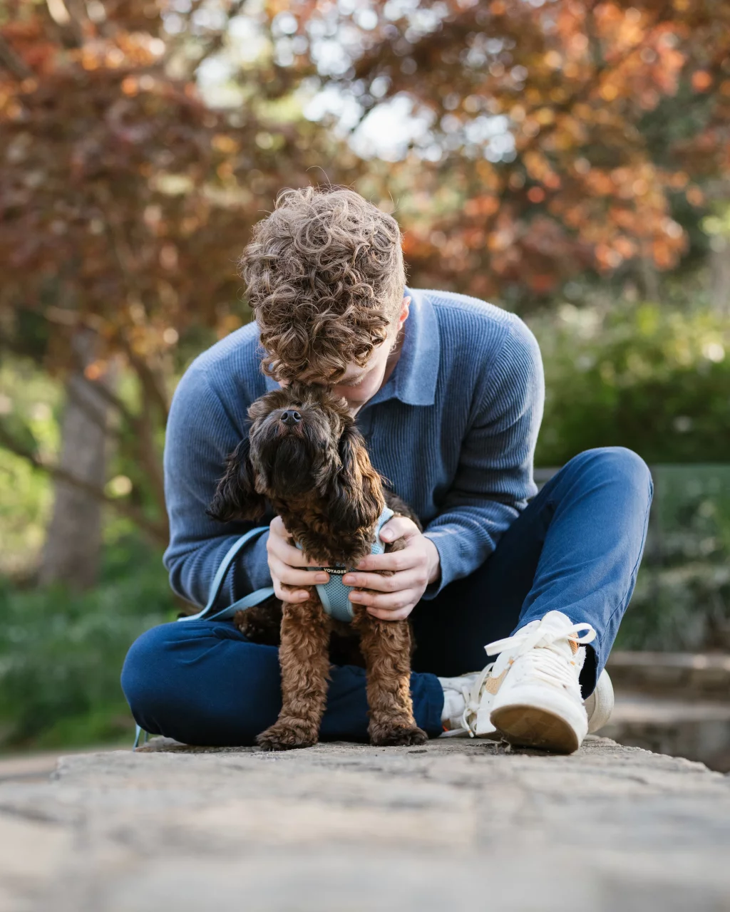 Jacobi's senior photos taken at Falls Park, Greenville SC: A person with curly hair sits cross-legged outdoors, gently holding and kissing a small dog wearing a harness. Autumn trees are blurred in the background.