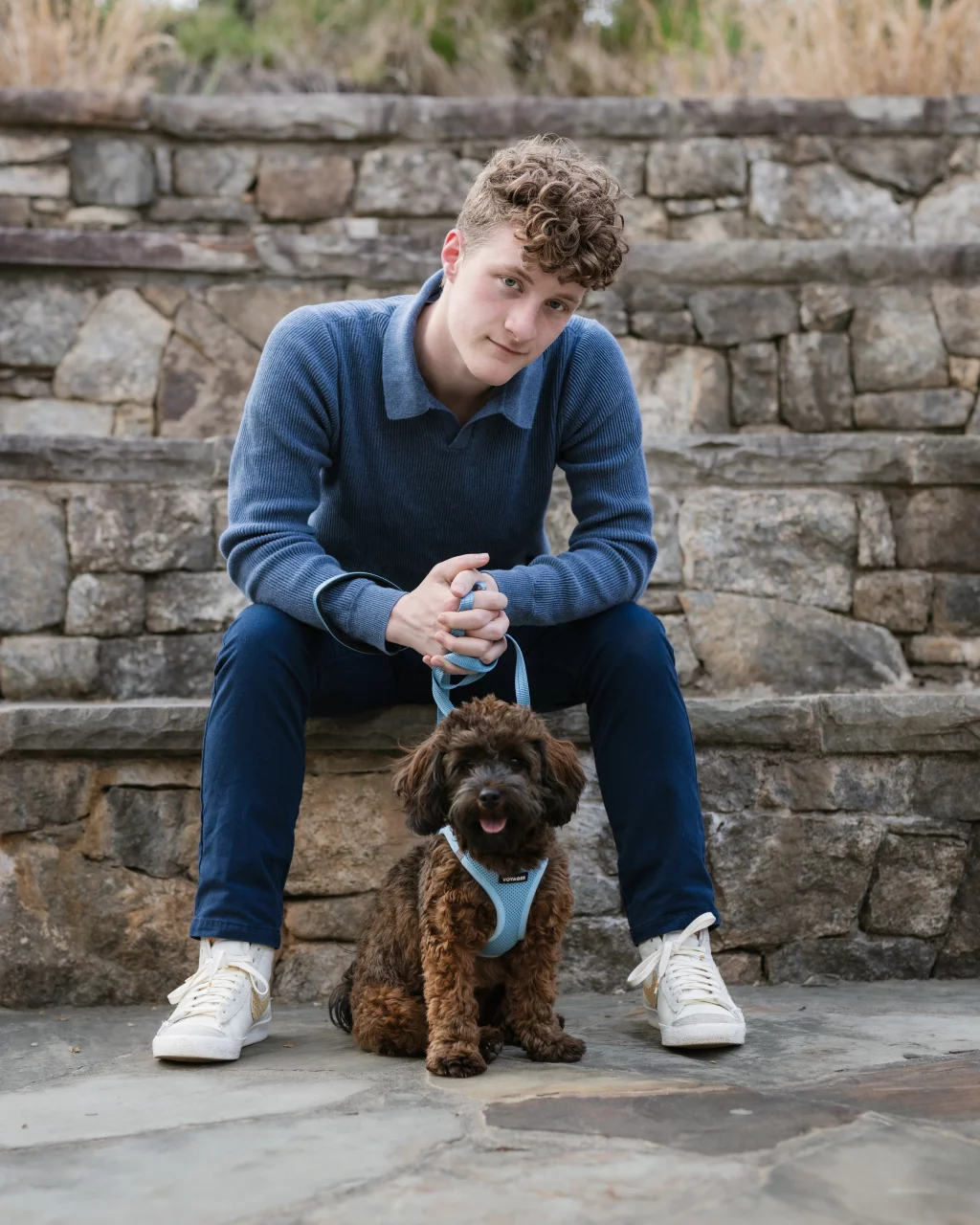 Jacobi's senior photos taken at Falls Park, Greenville SC: Young person with curly hair wearing a blue sweater and jeans sits on stone steps, holding a small brown dog on a blue harness.