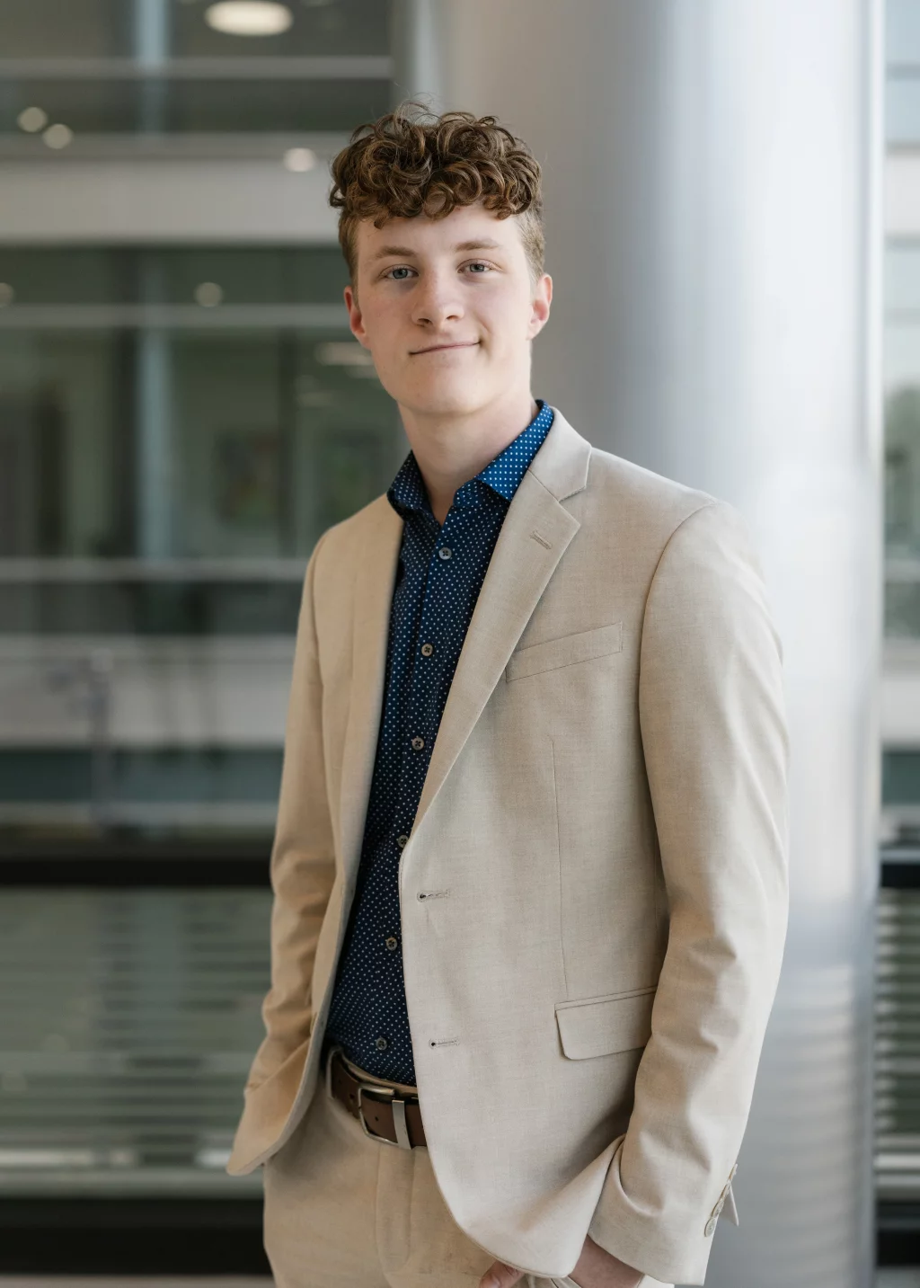 Jacobi's senior photos taken at Falls Park, Greenville SC: Young man with curly hair wearing a light beige suit and a navy blue patterned shirt, standing indoors in a modern building with glass and metal elements in the background.