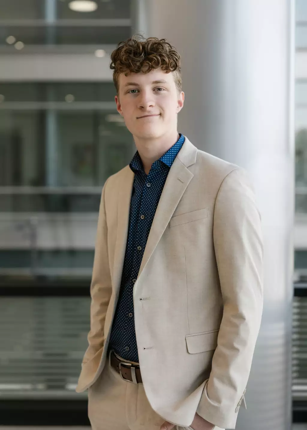 : Young man with curly hair wearing a light beige suit and a navy blue patterned shirt, standing indoors in a modern building with glass and metal elements in the background.
