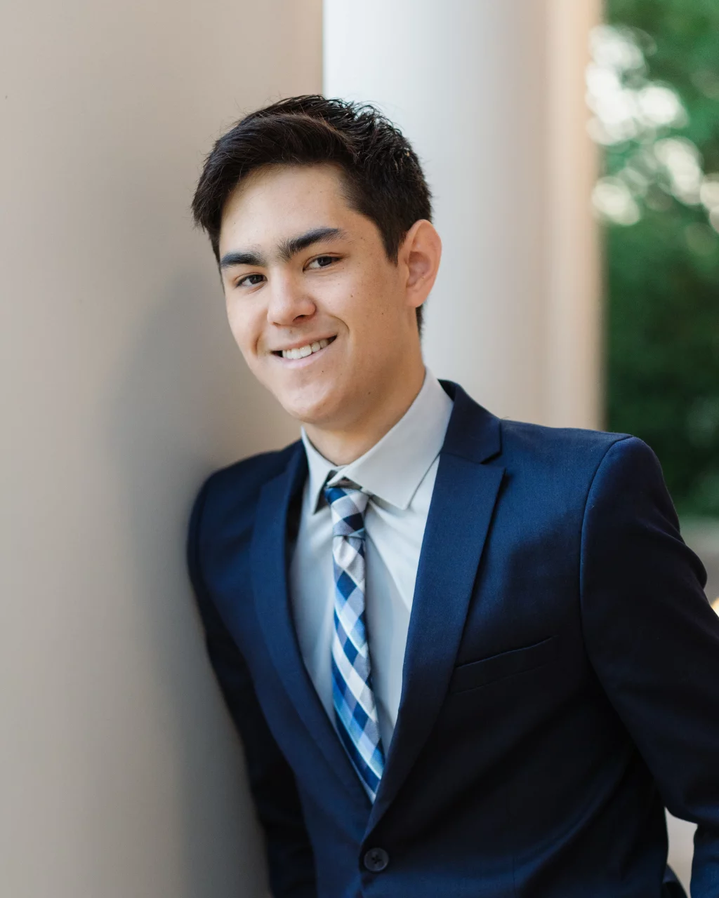 Jared's senior photos taken at Furman University: Young man in a navy suit and striped tie leaning against a light-colored wall, smiling at the camera.