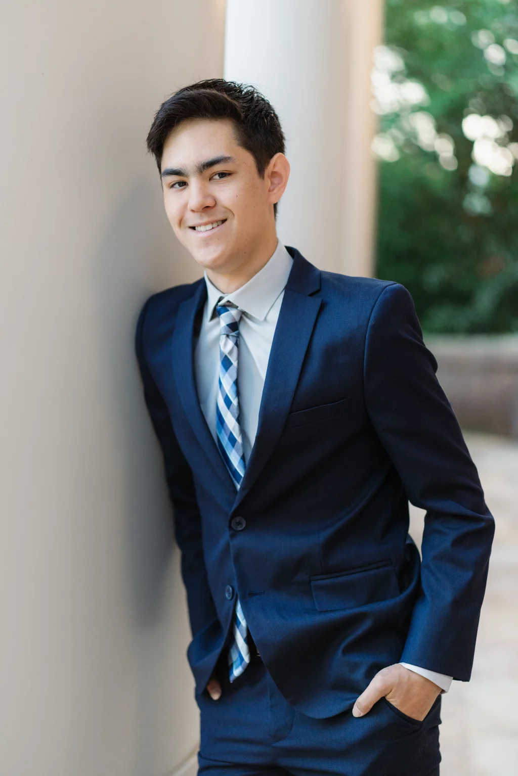 Jared's senior photos taken at Furman University: Young man in a blue suit and striped tie leaning against a wall with one hand in his pocket, smiling, with a blurred outdoor background.