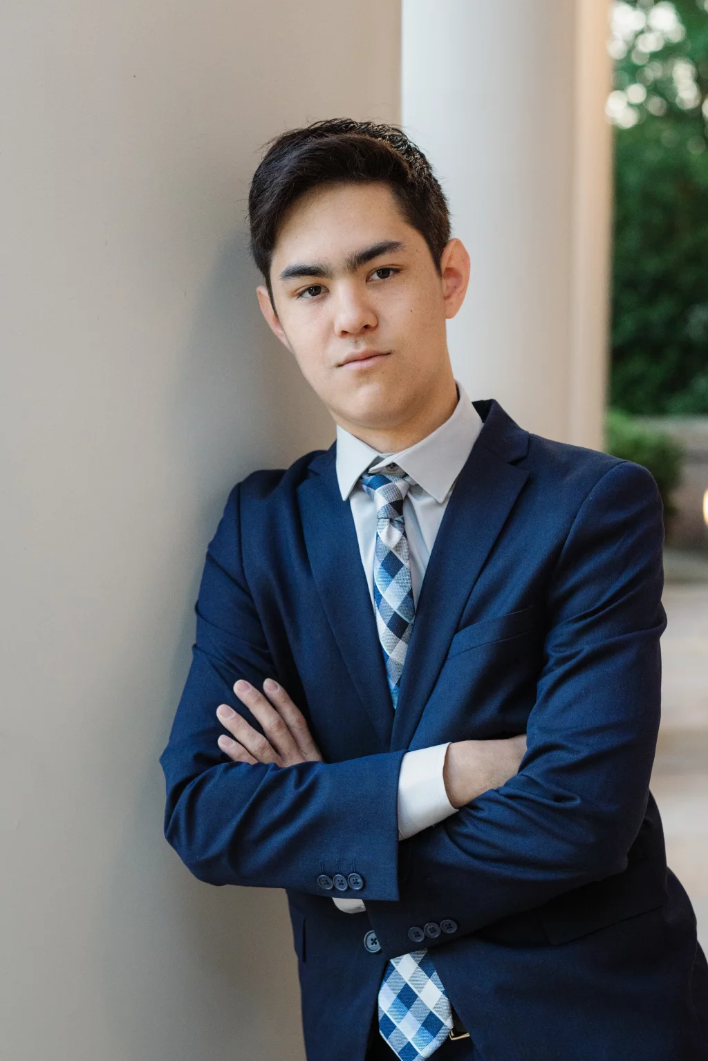 Jared's senior photos taken at Furman University: Young man in a blue suit and patterned tie standing with arms crossed, leaning against a light-colored wall, with greenery and columns in the background.