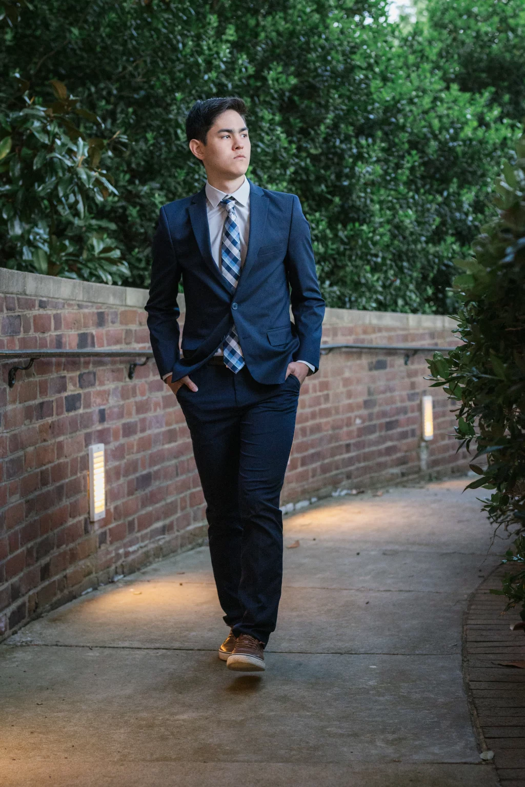 Jared's senior photos taken at Furman University: A young man in a navy suit and plaid tie walks along a curved brick pathway surrounded by greenery, with his hands in his pockets and looking to the side.