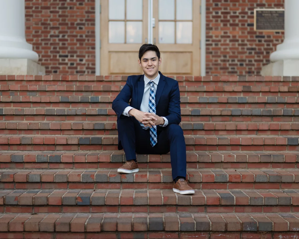 Jared's senior photos taken at Furman University: Young man in a navy suit and plaid tie sitting on brick steps in front of a building with double wooden doors and a brick wall.