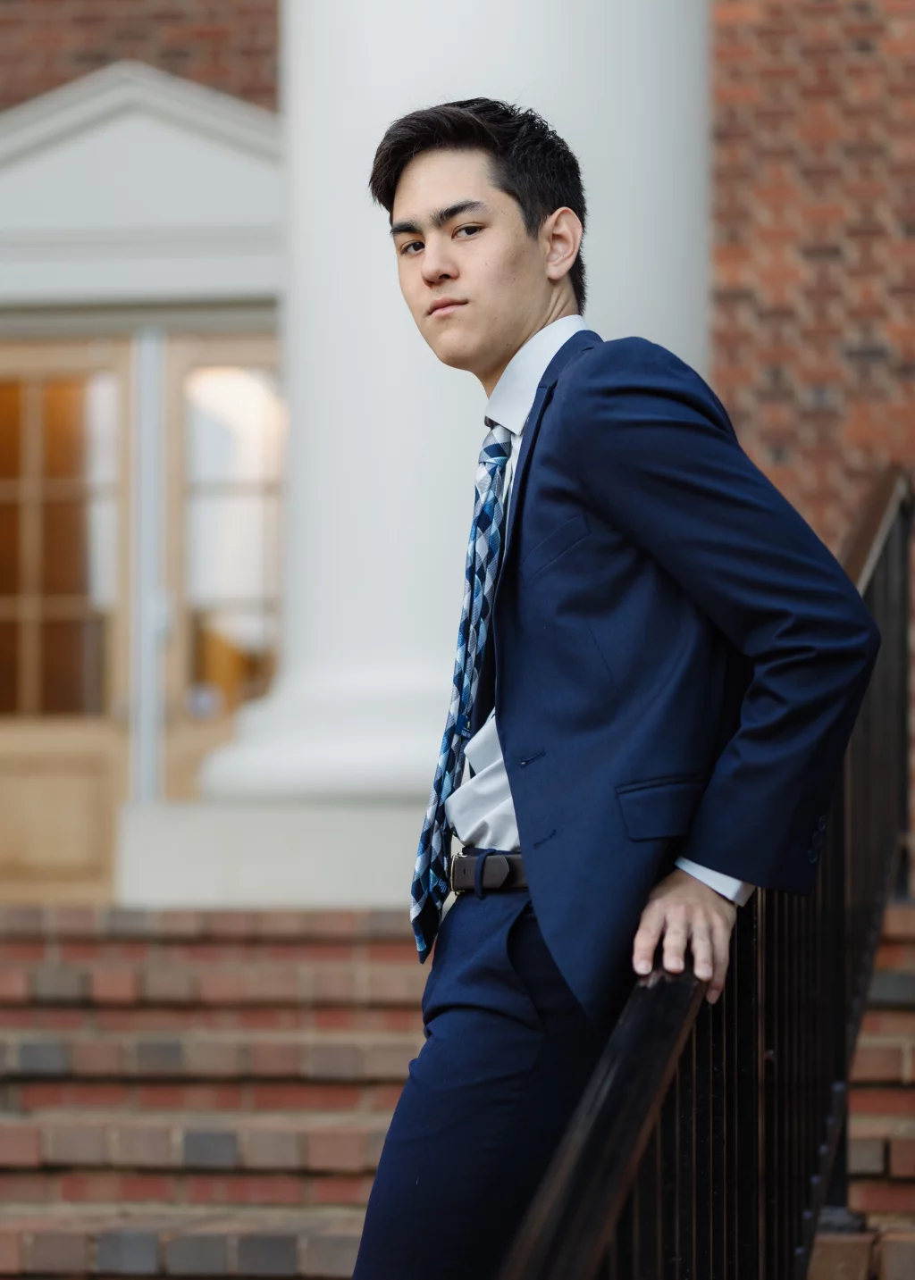 Jared's senior photos taken at Furman University: Young man in a blue suit and patterned tie leaning against a black railing on brick steps, with a building featuring white columns and large windows in the background.