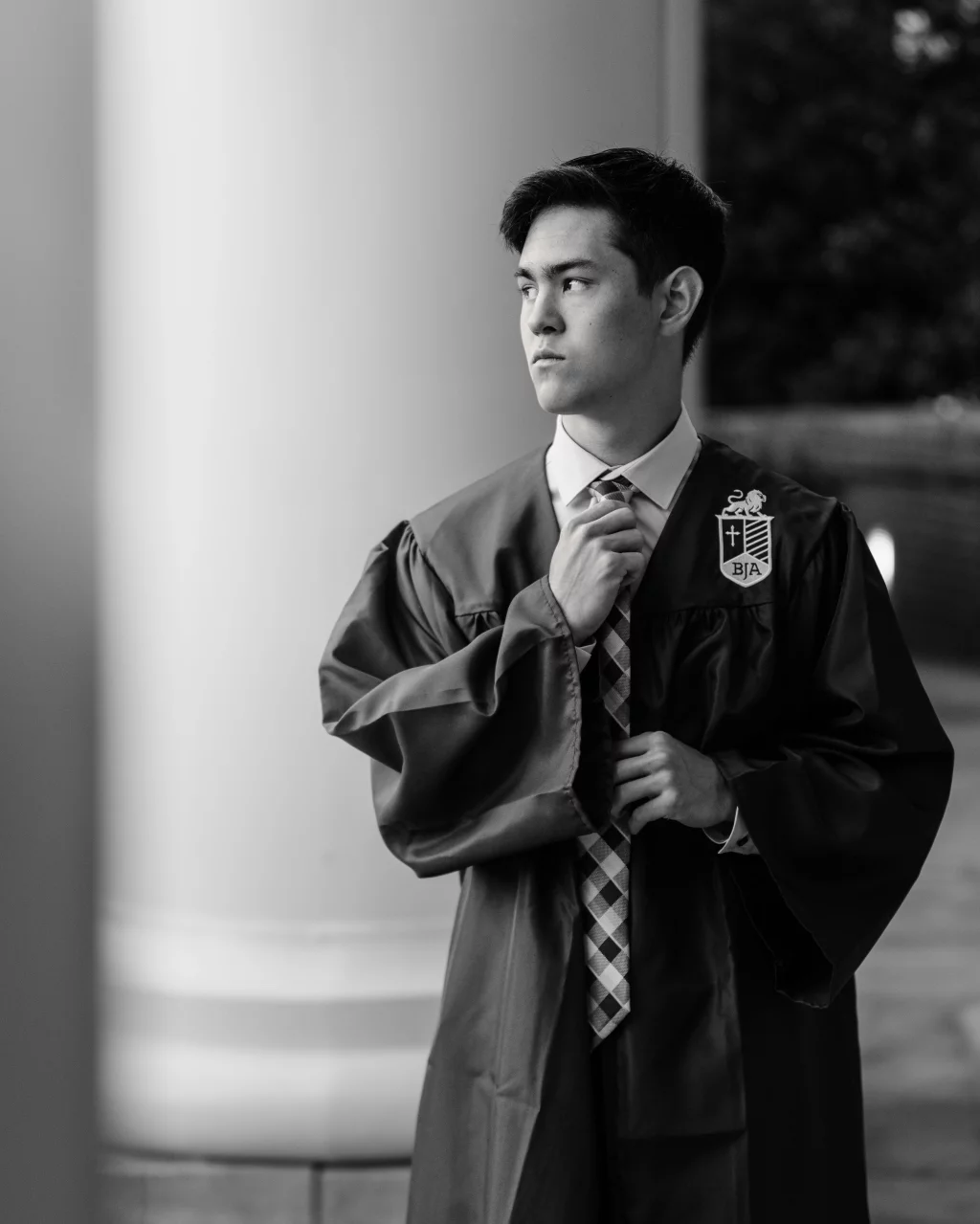 Jared's senior photos taken at Furman University: Black and white photo of a young man in a graduation gown adjusting his tie, standing near a large column and looking to the side.