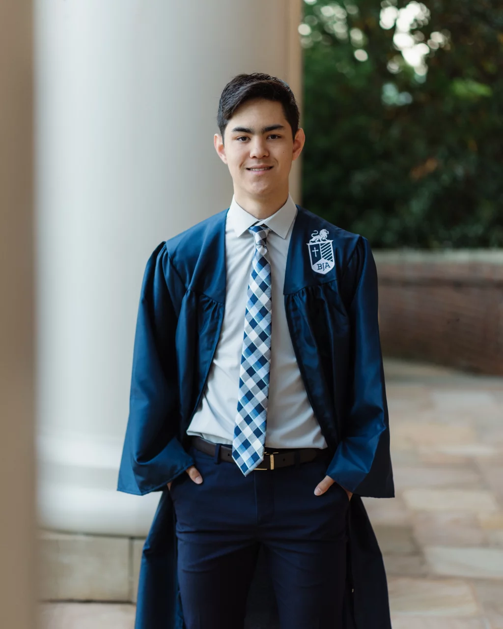 Jared's senior photos taken at Furman University: Young man wearing a blue graduation gown and a blue-and-white plaid tie, standing outdoors in front of a white column and greenery, with hands in his pockets.