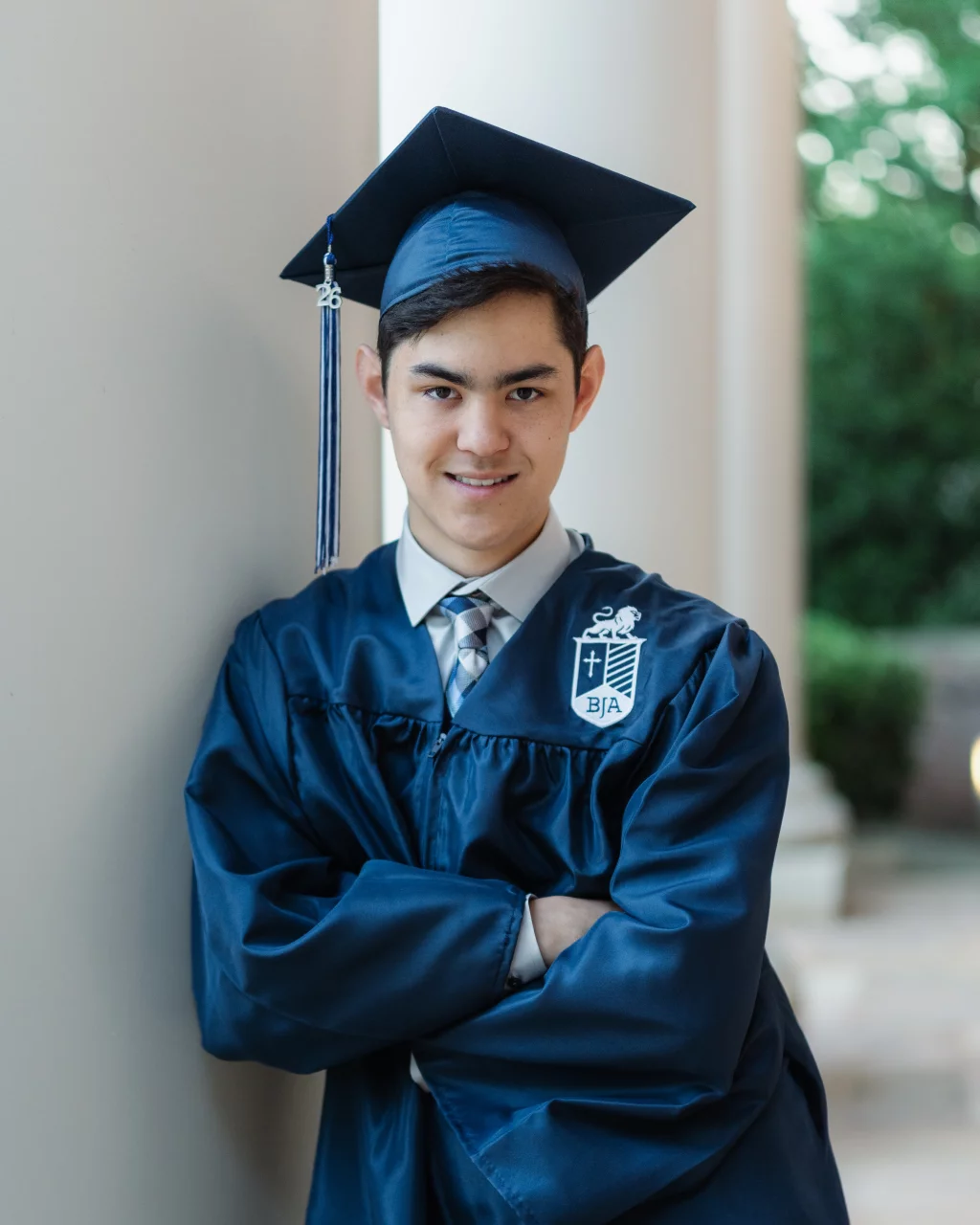 Jared's senior photos taken at Furman University: Young person in a blue graduation gown and cap with a tassel, standing with arms crossed and smiling, leaning against a white column.