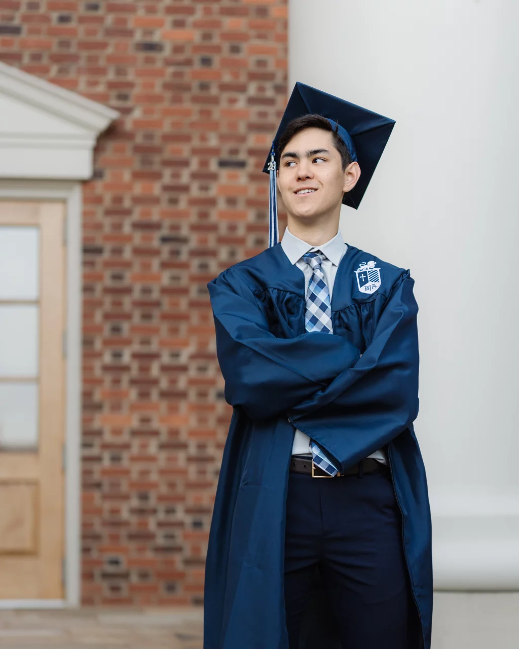 Jared's senior photos taken at Furman University: Young man wearing a blue graduation cap and gown with arms crossed, standing in front of a brick building.