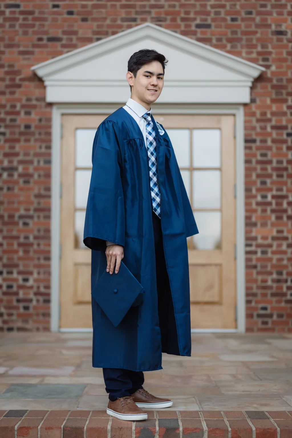 Jared's senior photos taken at Furman University: A young man wearing a blue graduation gown and checkered tie stands in front of a brick building, holding a matching blue graduation cap at his side.