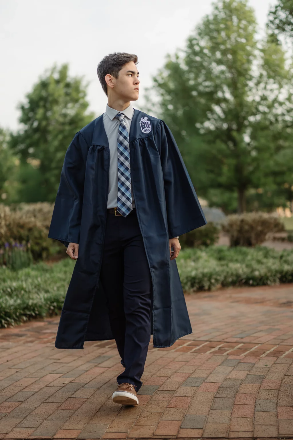 Jared's senior photos taken at Furman University: A young man in a graduation gown and tie walks outdoors on a brick path, with greenery and trees in the background.