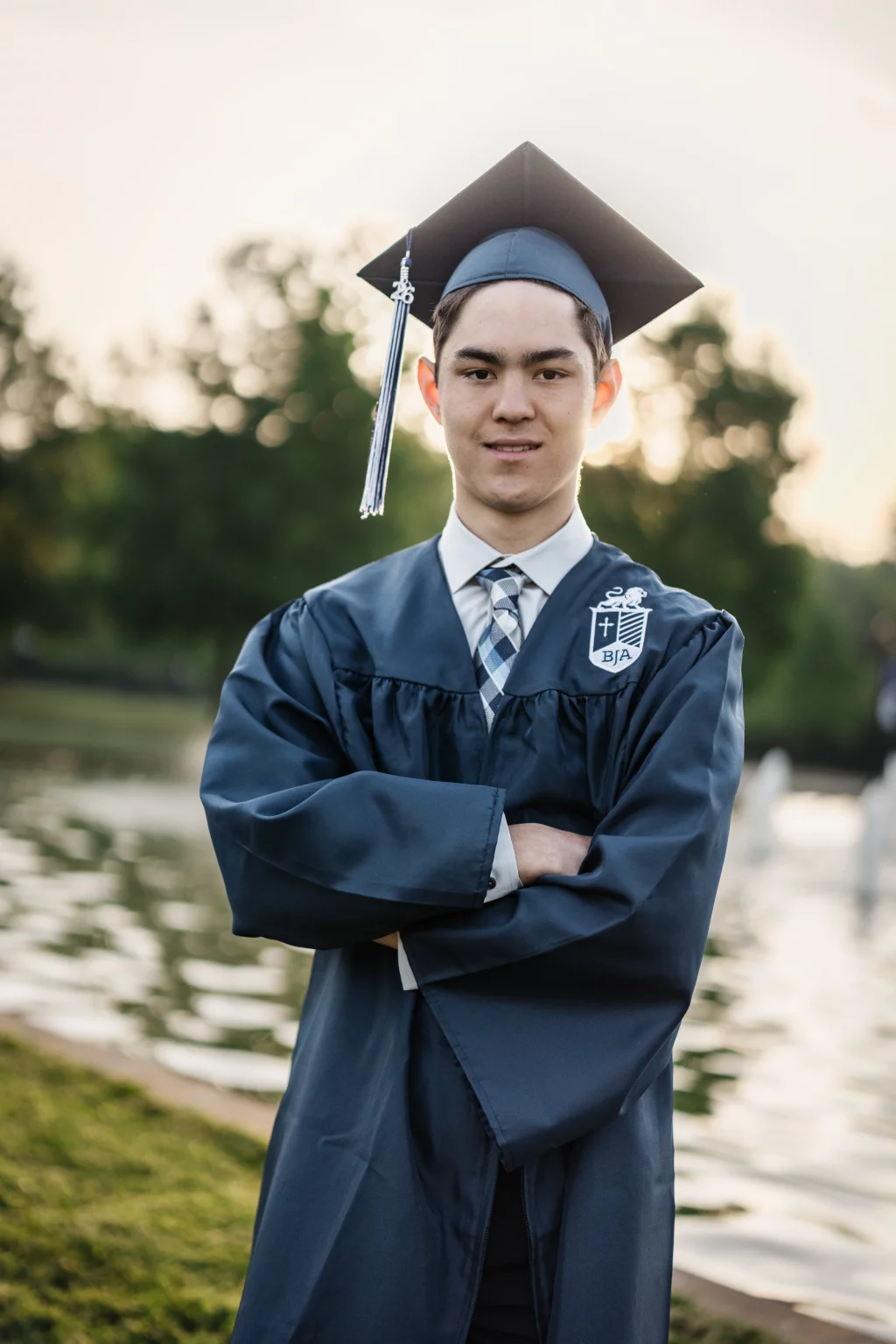 Jared's senior photos taken at Furman University: Young man wearing a blue graduation cap and gown with arms crossed, standing outdoors near a pond with trees in the background.