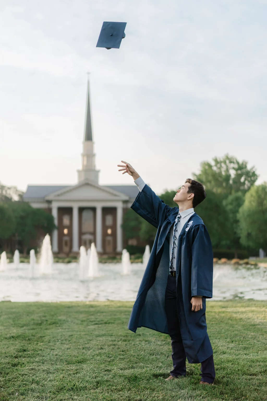 Jared's senior photos taken at Furman University: A graduate in a blue cap and gown stands on grass in front of a fountain and brick building with a tall steeple, tossing their graduation cap into the air.