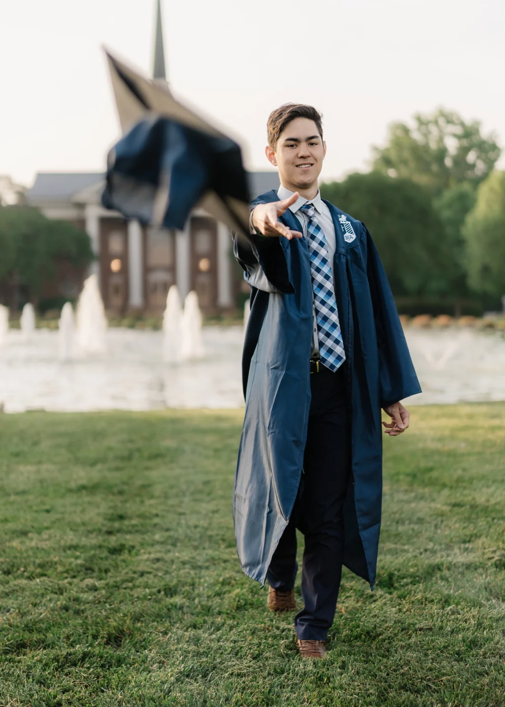 Jared's senior photos taken at Furman University: Young man in a blue graduation gown and plaid tie tossing his cap, walking on grass with a fountain and academic building in the background.