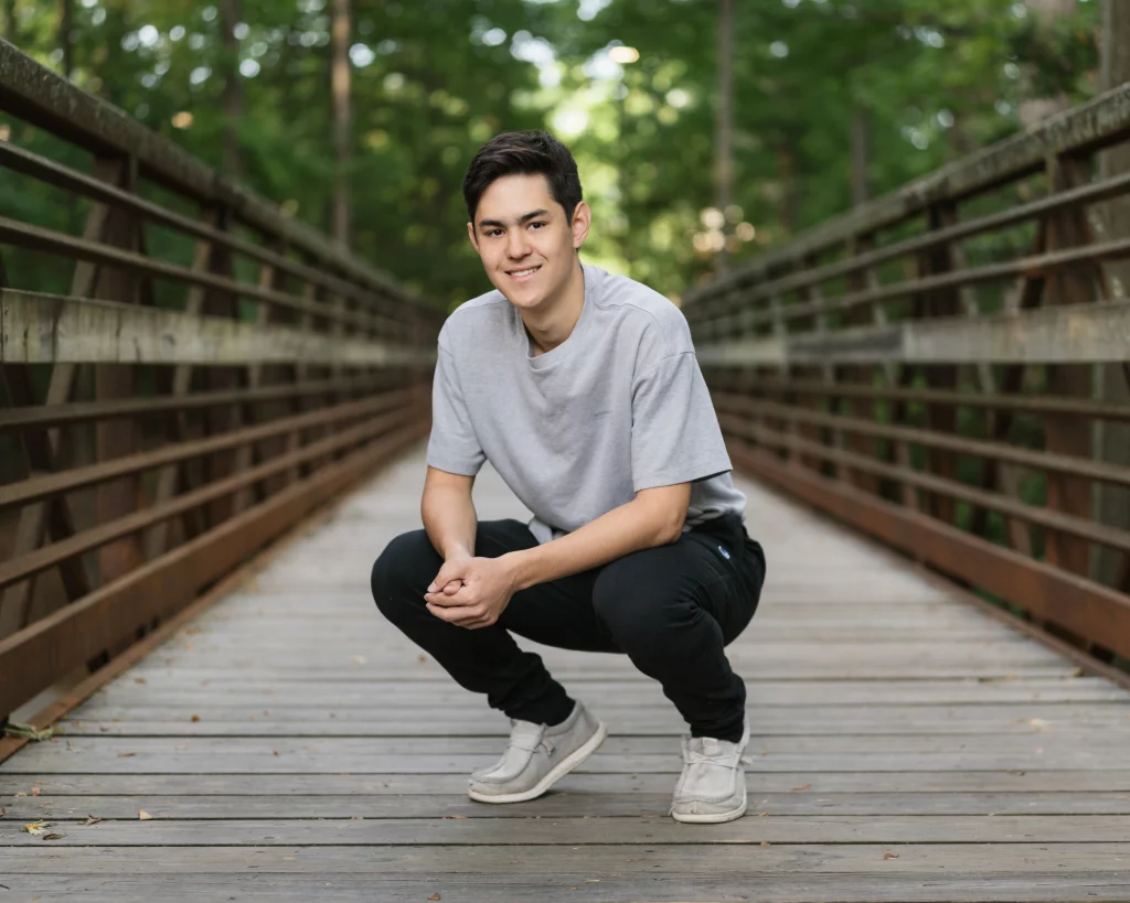 Jared's senior photos taken at Furman University: A young man squats and smiles on a wooden bridge surrounded by trees.
