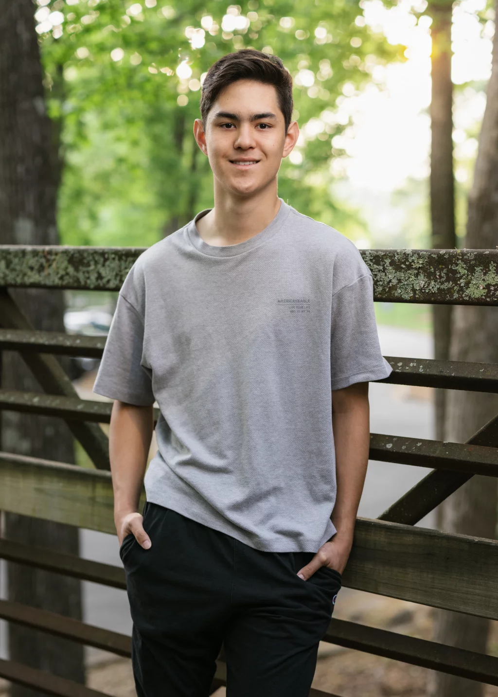 Jared's senior photos taken at Furman University: Young man standing outdoors, leaning against a wooden and metal fence, wearing a gray t-shirt and black pants, with trees and sunlight in the background.