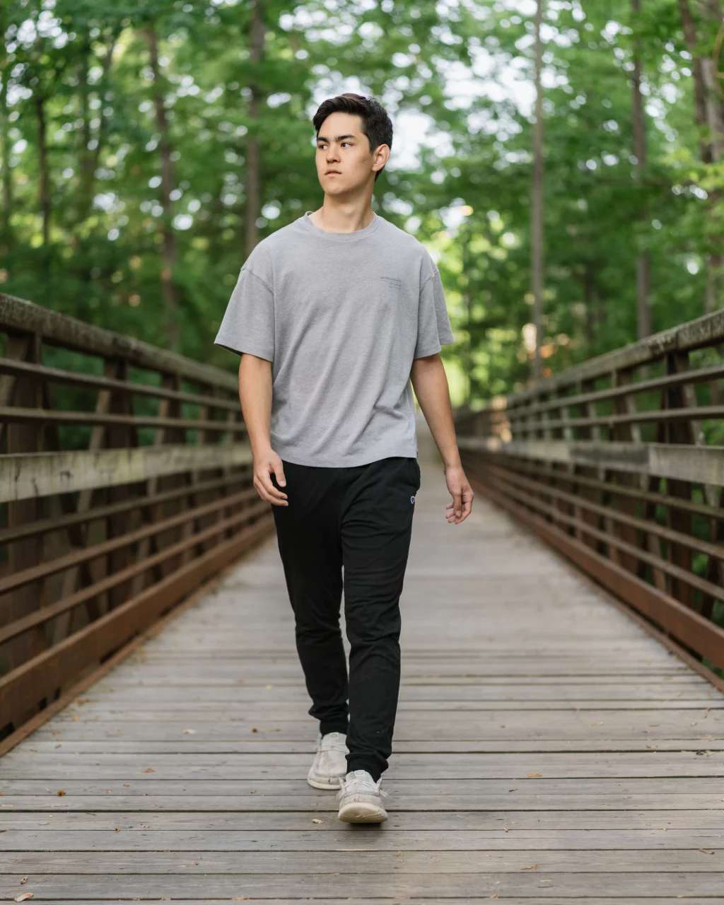 Jared's senior photos taken at Furman University: A young man wearing a gray t-shirt, black pants, and white sneakers walks on a wooden bridge surrounded by green trees.