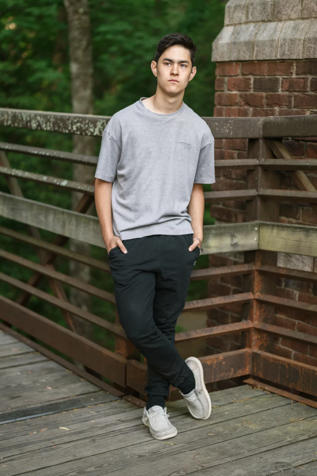 Jared's senior photos taken at Furman University: A young person with short dark hair, wearing a gray t-shirt, black pants, and gray slip-on shoes, stands with hands in pockets and legs crossed on a wooden bridge with a brick column and metal railing, surrounded by greenery.