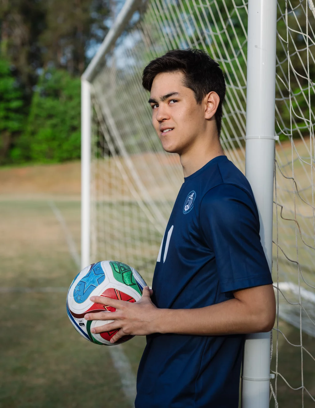 Jared's senior photos taken at Furman University: Young soccer player in a navy blue jersey holding a soccer ball, leaning against a goalpost on an outdoor field.