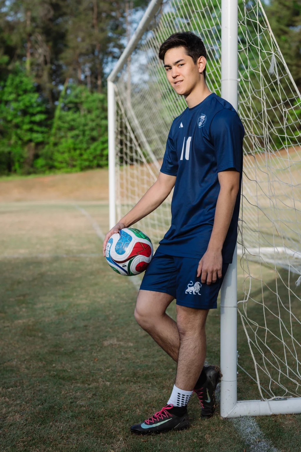 Jared's senior photos taken at Furman University: Young male soccer player in a blue uniform leaning against a goalpost, holding a soccer ball on a grassy field with trees in the background.