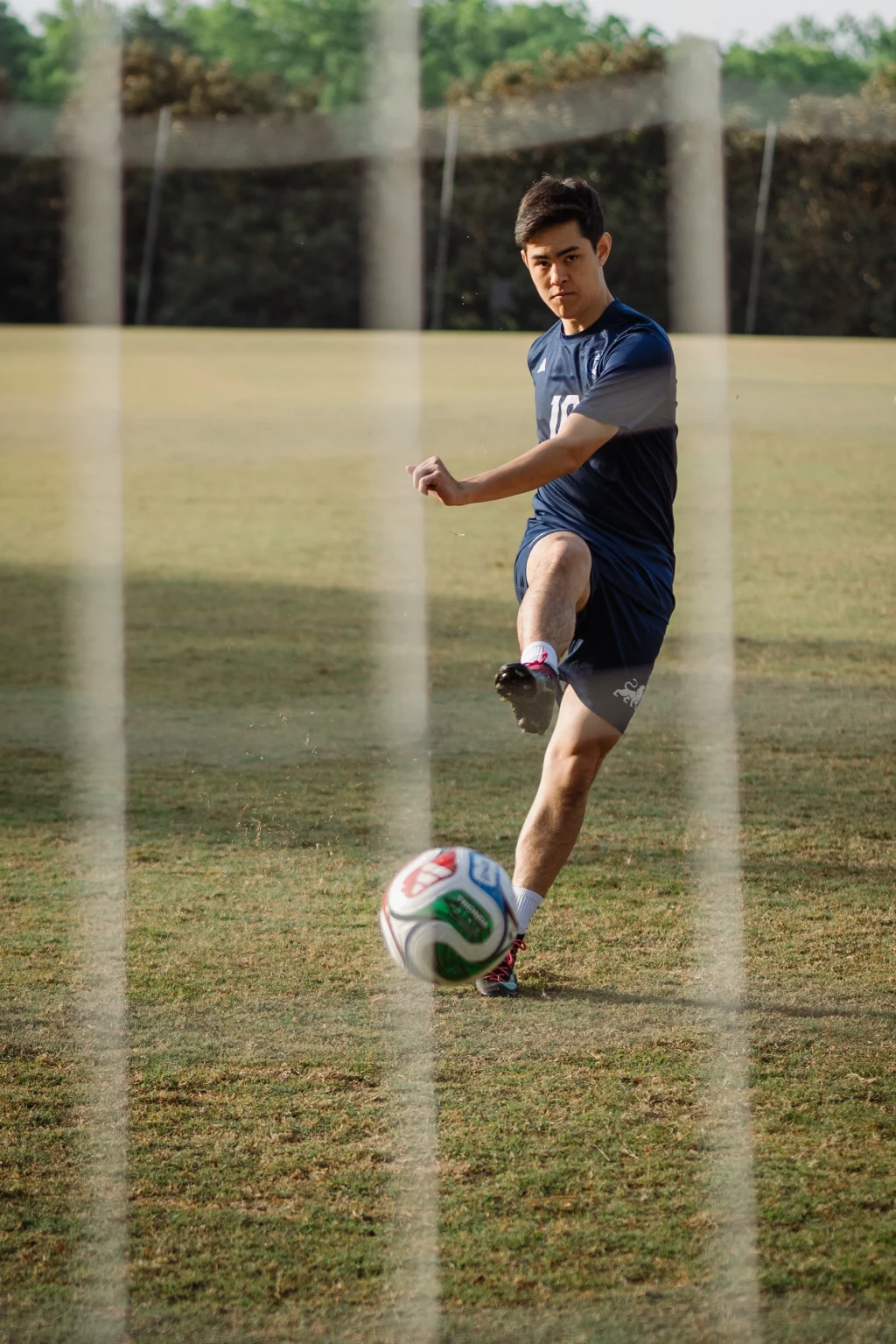 Jared's senior photos taken at Furman University: A soccer player in a navy blue uniform is kicking a soccer ball on a grassy field, viewed through the netting of a goal.