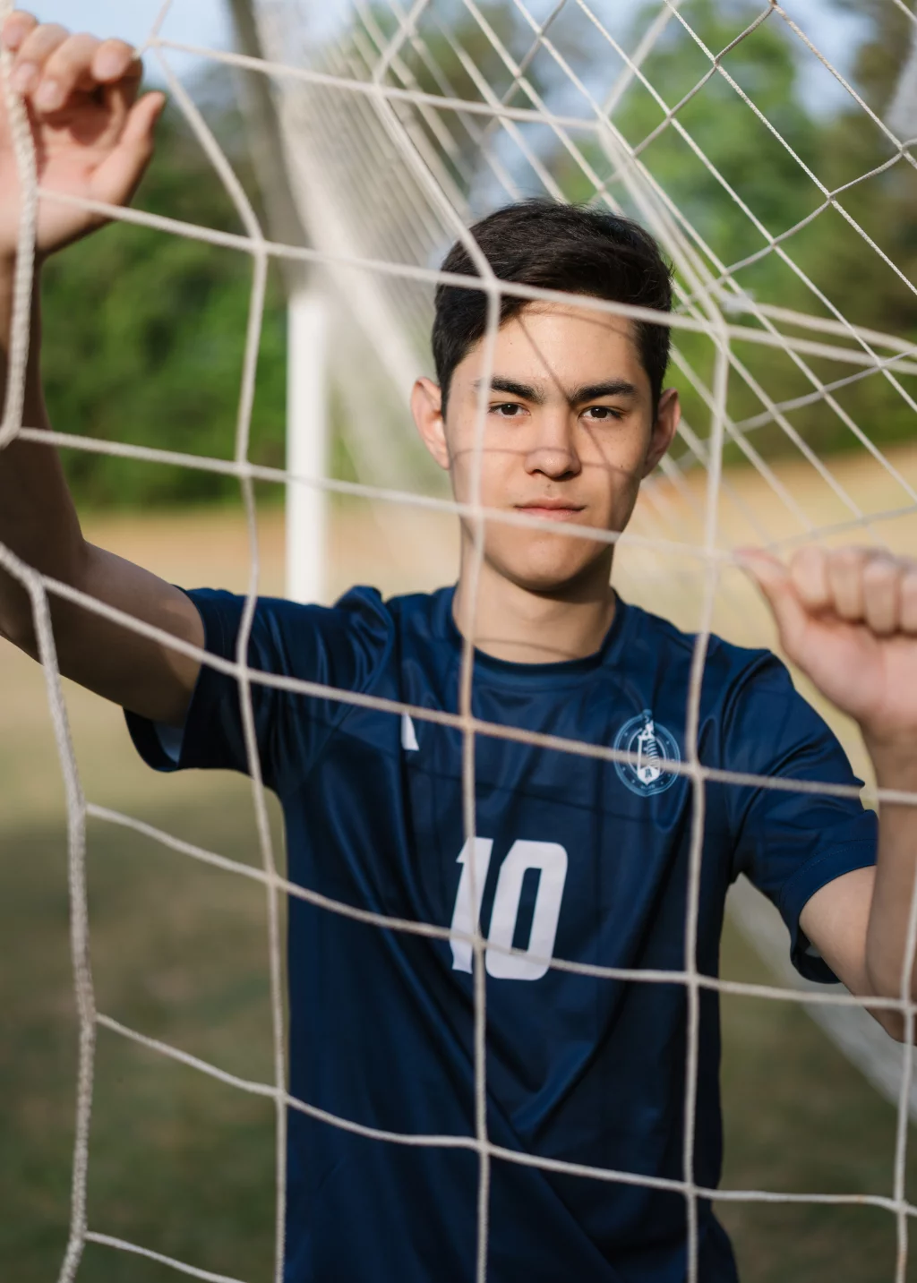 Jared's senior photos taken at Furman University: Young soccer player in a blue jersey with the number 10 stands behind a goal net, holding onto the net with both hands and looking forward.