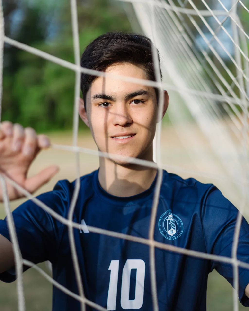 Jared's senior photos taken at Furman University: A young soccer player in a blue jersey with the number 10 stands behind a soccer goal net, looking toward the camera with hands touching the net.