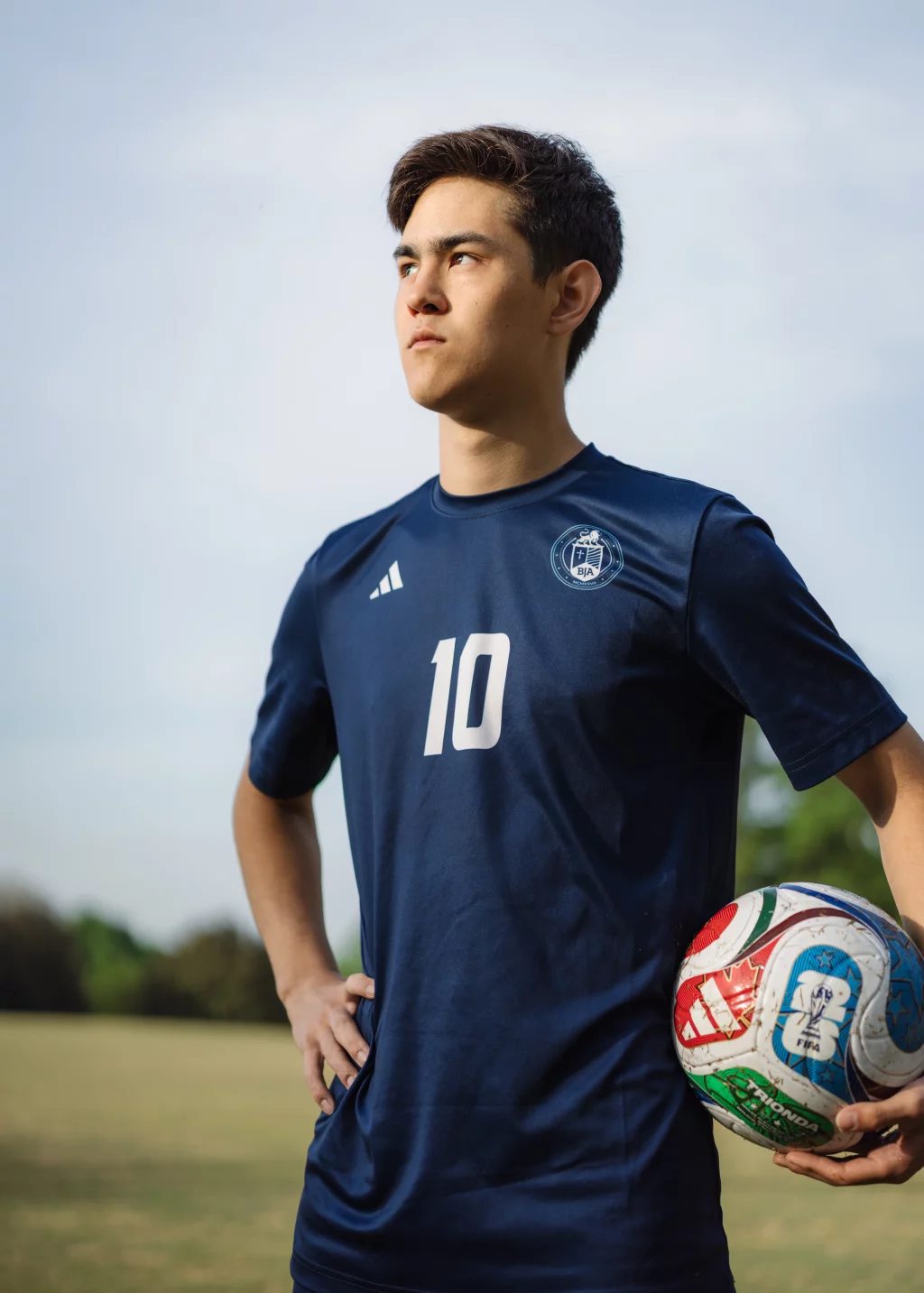 Jared's senior photos taken at Furman University: Young soccer player in a navy blue jersey with the number 10 stands on a field holding a colorful soccer ball under one arm, looking determinedly into the distance.