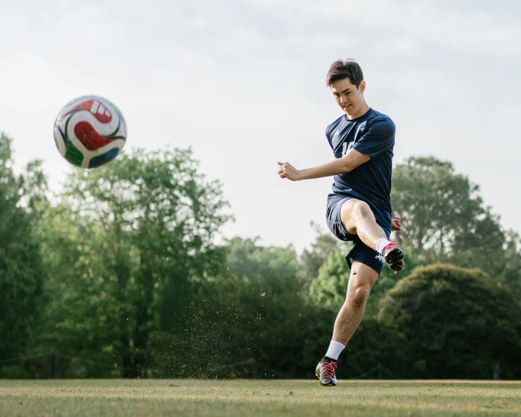 Jared's senior photos taken at Furman University: A soccer player in a navy uniform kicks a soccer ball on a grassy field with trees in the background.