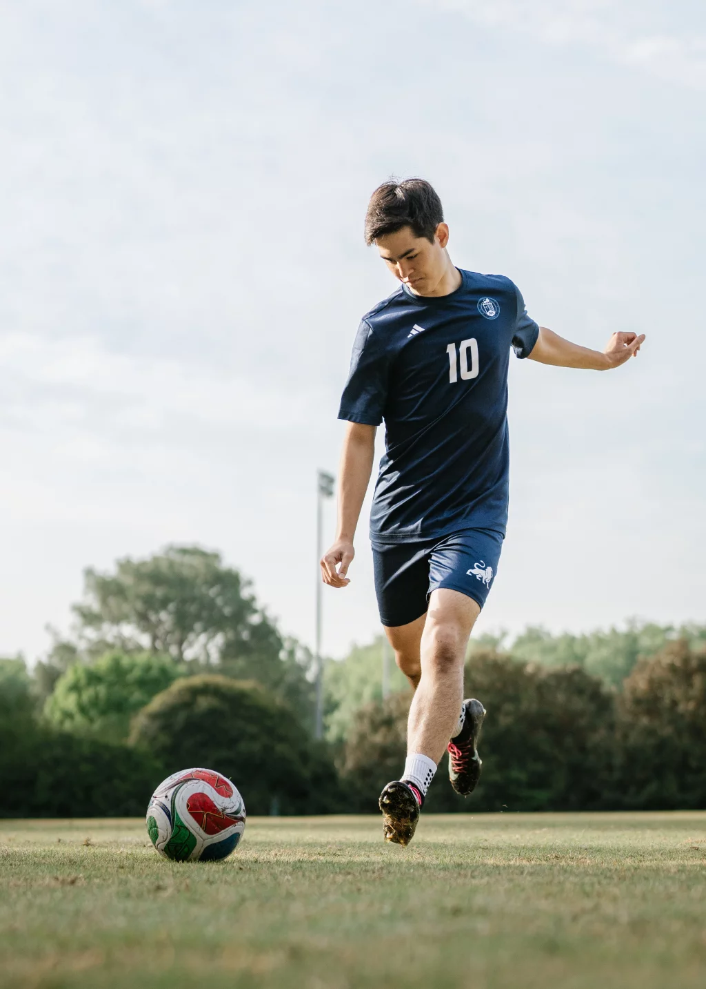 Jared's senior photos taken at Furman University: A soccer player in a navy blue uniform with the number 10 prepares to kick a soccer ball on a grassy field, with trees and a light post in the background.