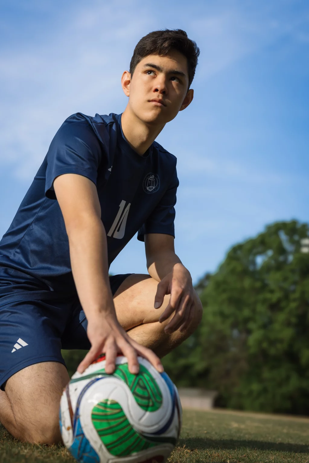 Jared's senior photos taken at Furman University: A young soccer player in a navy blue uniform kneels on a grassy field, resting one hand on a soccer ball, with trees and blue sky in the background.