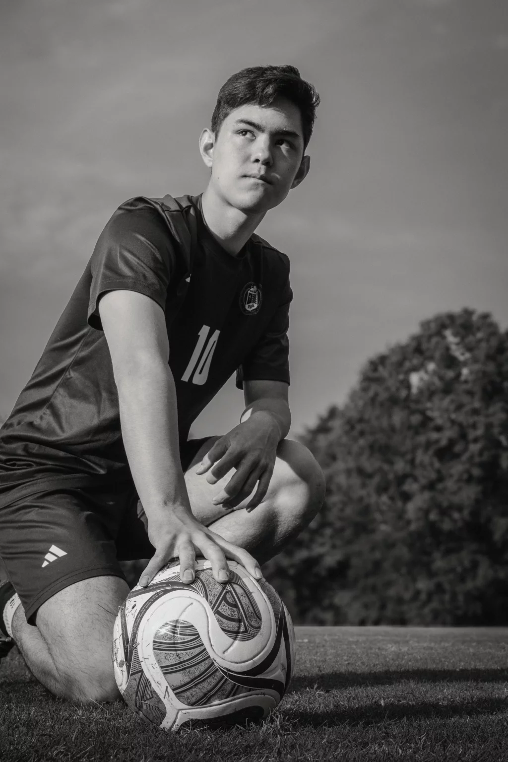Jared's senior photos taken at Furman University: Black and white photo of a young male soccer player kneeling on grass with one hand resting on a soccer ball, looking off into the distance. Trees are visible in the background.
