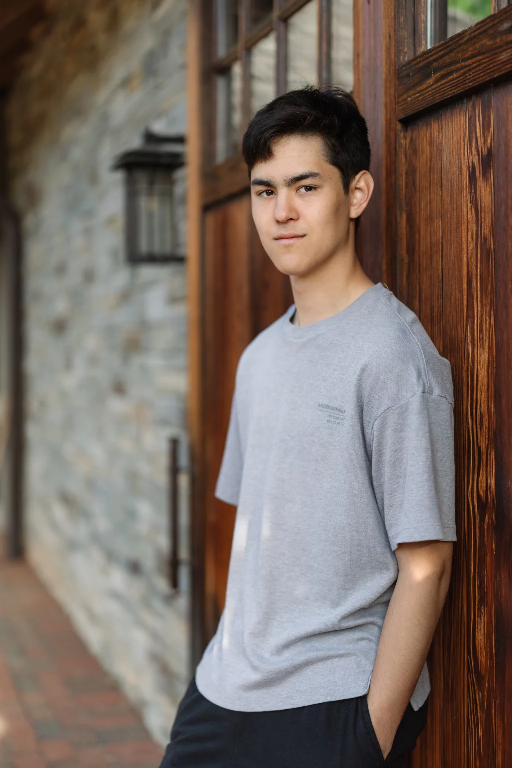 Jared's senior photos taken at Furman University: Teen boy with short dark hair wearing a gray t-shirt and black pants, standing with hands in pockets and leaning against a wooden door, with a stone wall and lantern in the background.
