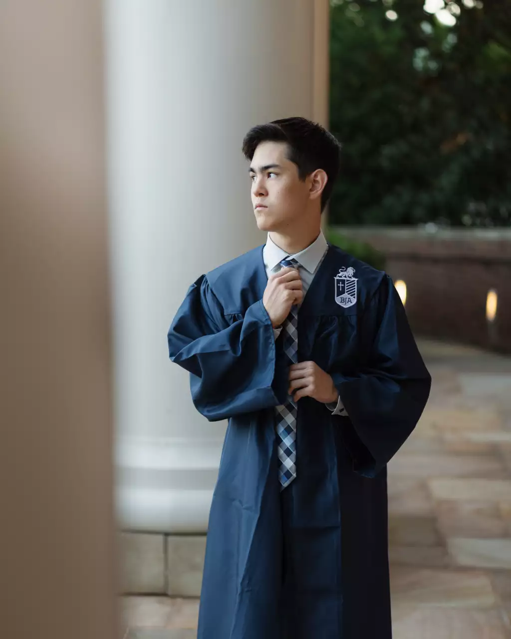 Jared's senior photos taken at Furman University: A young man in a blue graduation gown adjusts his tie while standing outdoors near a large white column, looking thoughtfully to the side.