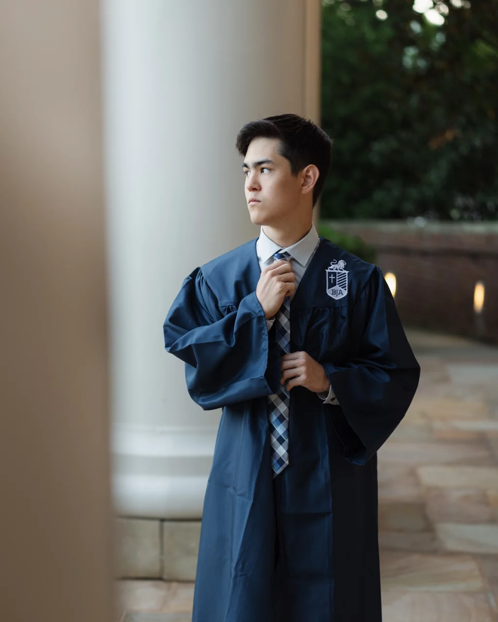 Jared's senior photos taken at Furman University: A young man in a blue graduation gown adjusts his tie while standing outdoors near a large white column, looking thoughtfully to the side.