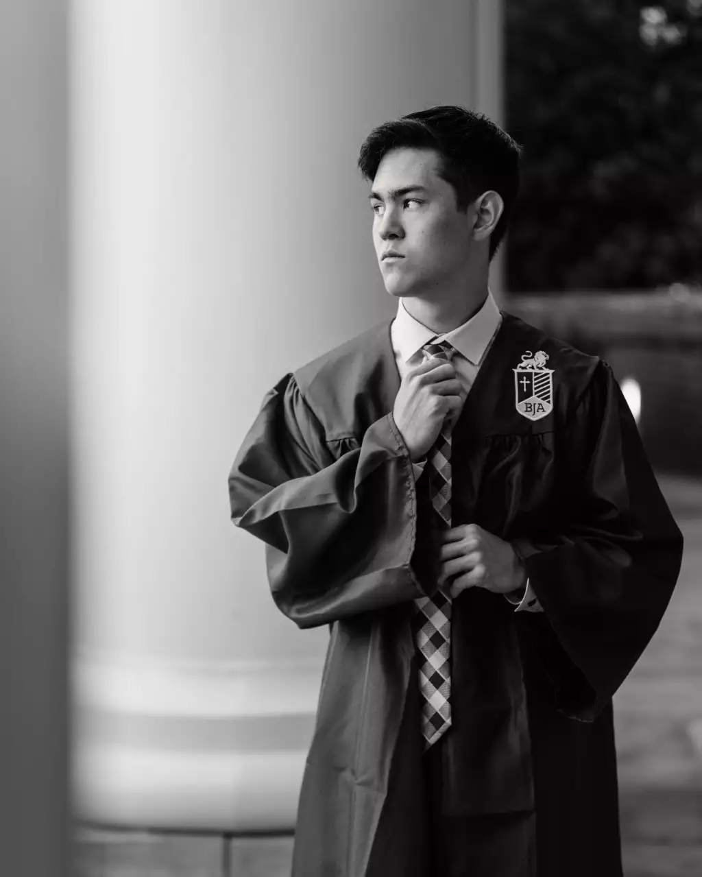 : Black and white photo of a young man in a graduation gown adjusting his tie, looking to the side, standing in front of a large column.
