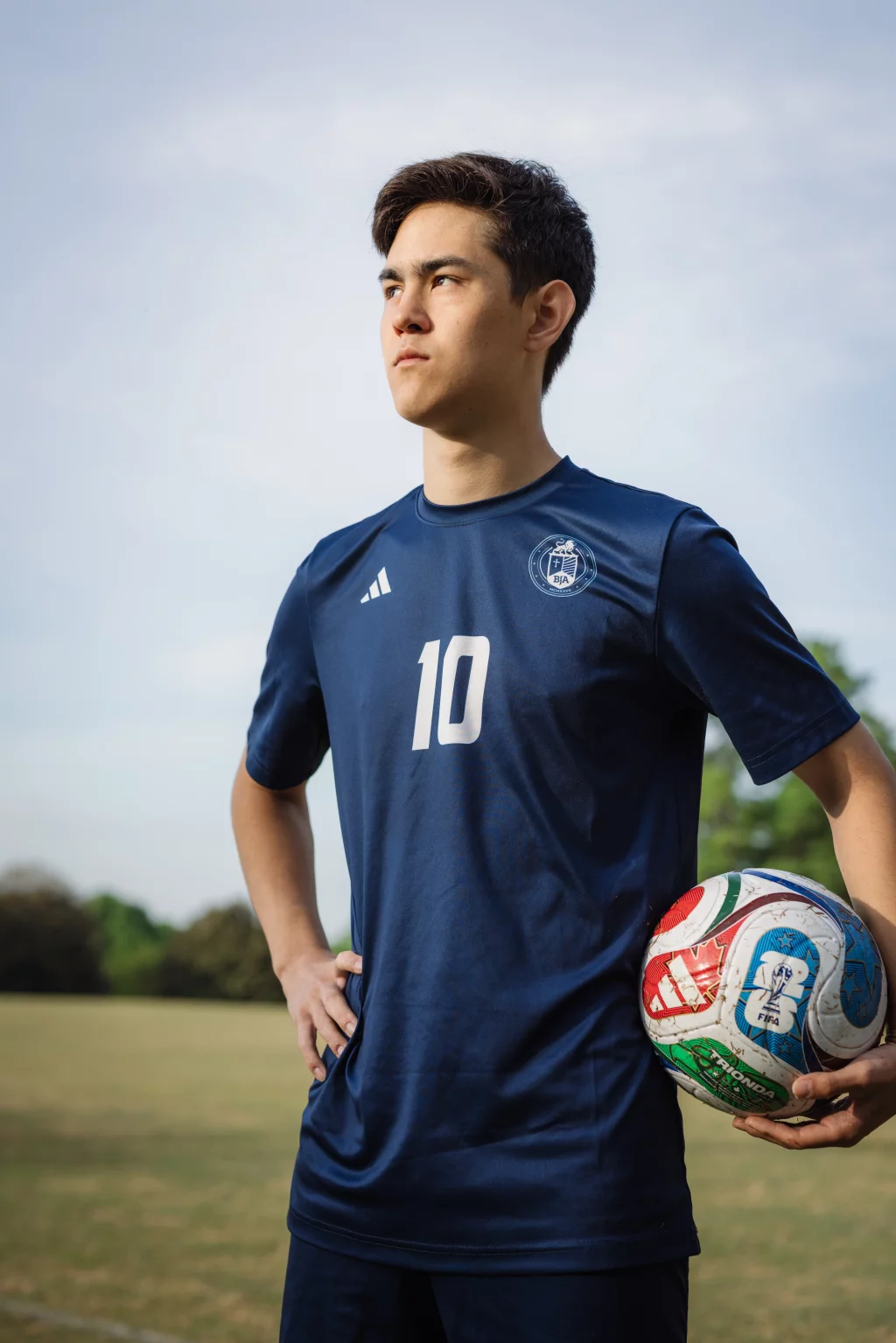 Jared's senior photos taken at Furman University: Young soccer player in a navy blue jersey with the number 10, standing on a field holding a soccer ball under one arm, looking determinedly into the distance.