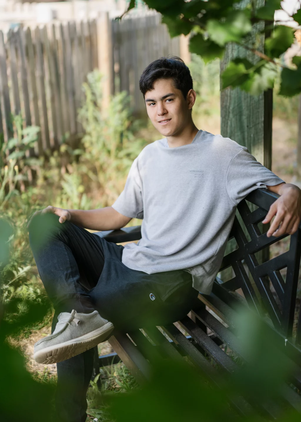 Jared's senior photos taken at Furman University: Young man sitting on a wooden bench outdoors, surrounded by greenery and a wooden fence, looking at the camera with a relaxed expression.