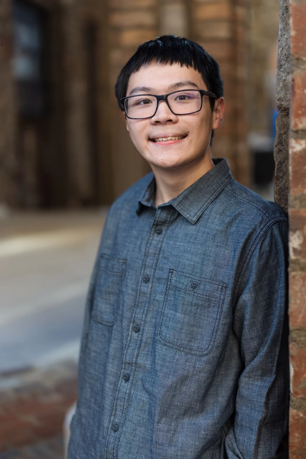 Johnny's senior photos taken at BridgeWay Station, Simpsonville, SC: A young person with short dark hair and glasses, wearing a blue button-up shirt, smiles while leaning against a brick wall in an outdoor setting.