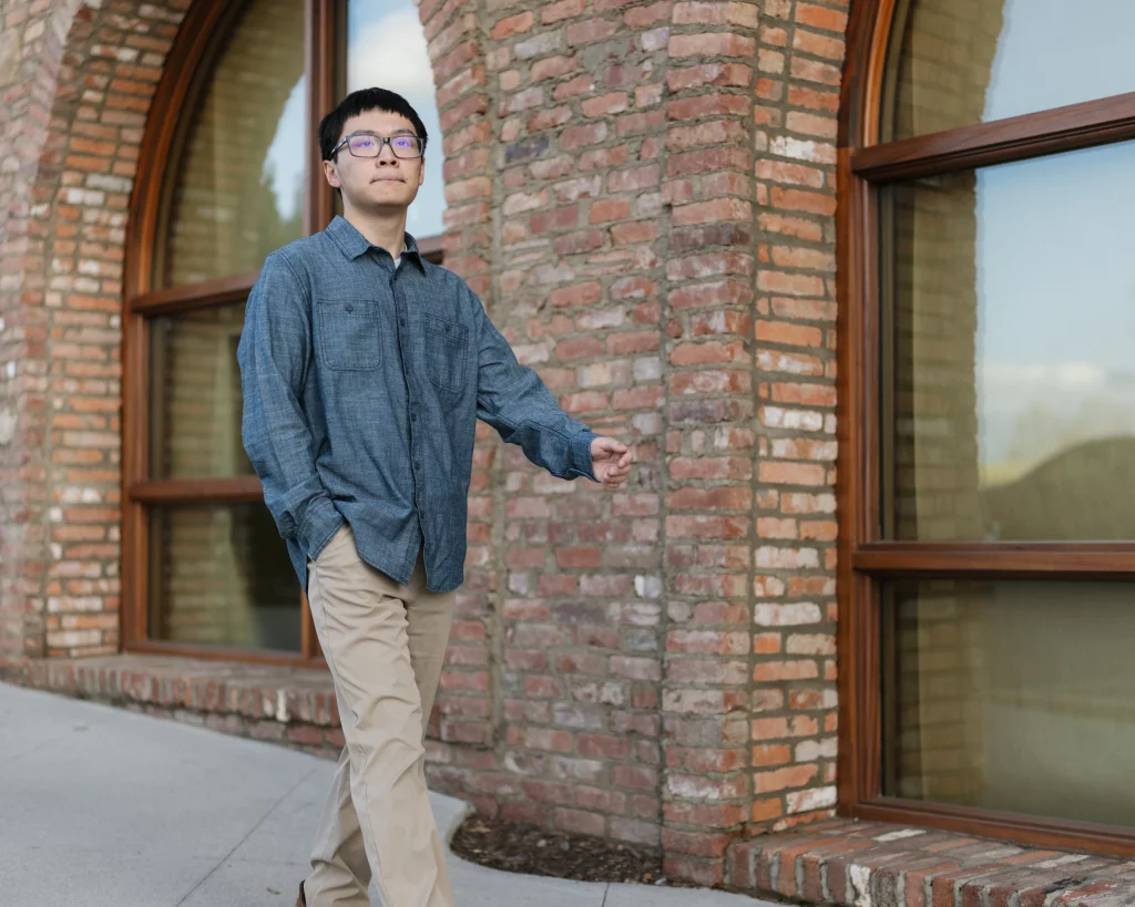 Johnny's senior photos taken at BridgeWay Station, Simpsonville, SC: Young man wearing glasses, a blue button-up shirt, and khaki pants walks on a sidewalk in front of a brick building with large arched windows.