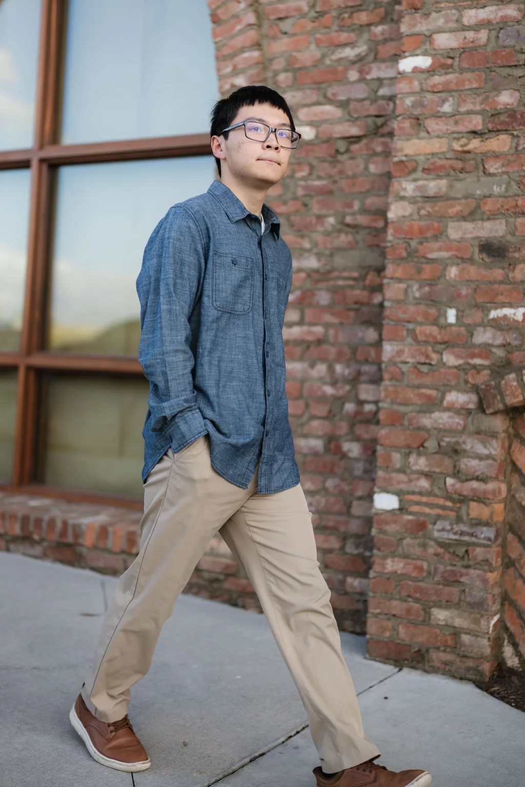 Johnny's senior photos taken at BridgeWay Station, Simpsonville, SC: A young man wearing glasses, a blue button-up shirt, khaki pants, and brown shoes walks outside past a brick wall and large window.