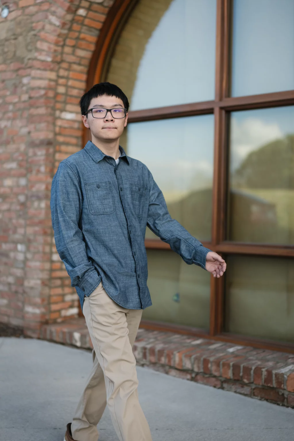 Johnny's senior photos taken at BridgeWay Station, Simpsonville, SC: A young man wearing glasses, a blue button-up shirt, and khaki pants walks outside in front of a large arched window set in a brick wall.