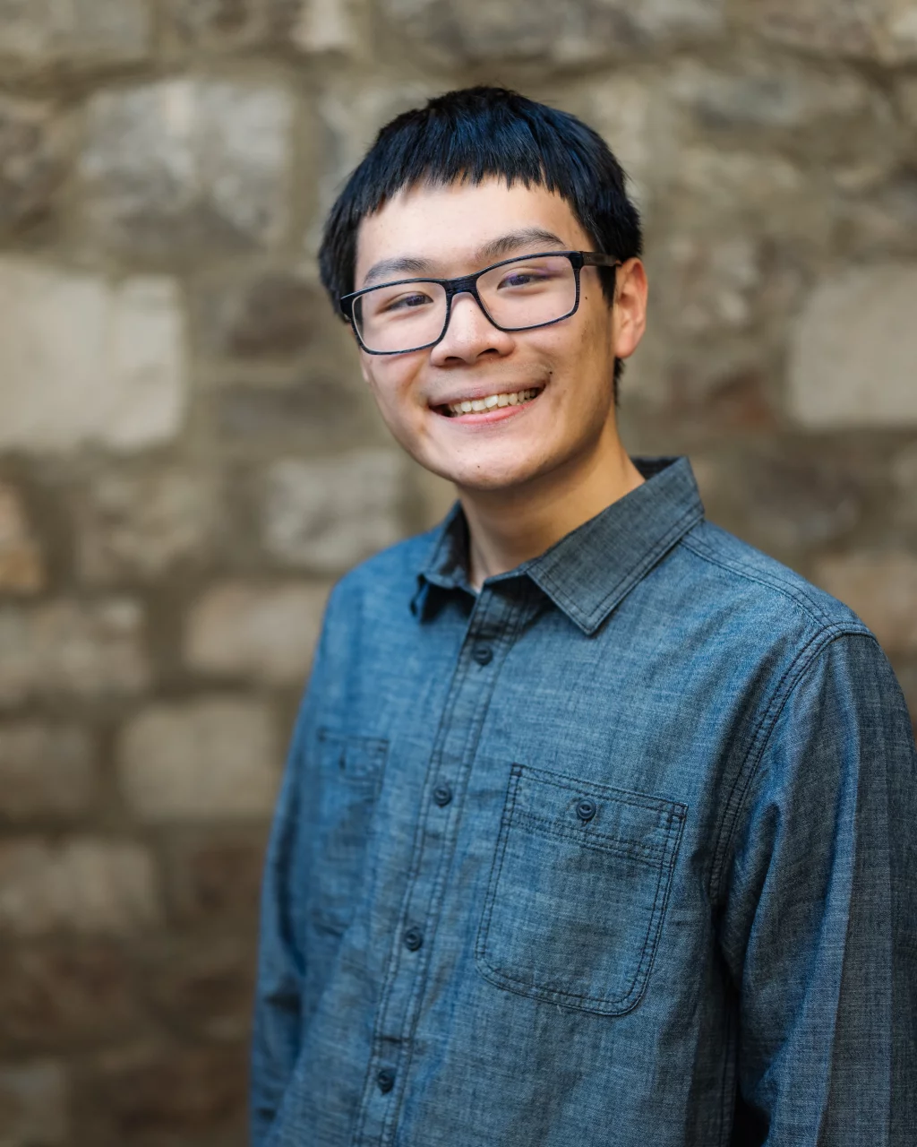 Johnny's senior photos taken at BridgeWay Station, Simpsonville, SC: Young person with short dark hair and glasses, smiling and wearing a blue button-up shirt, standing in front of a stone wall.